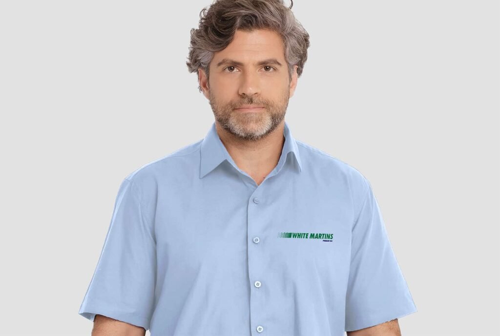 Homem vestindo camisa social masculina azul claro de manga curta com bordado White Martins aplicado no lado esquerdo do peito.