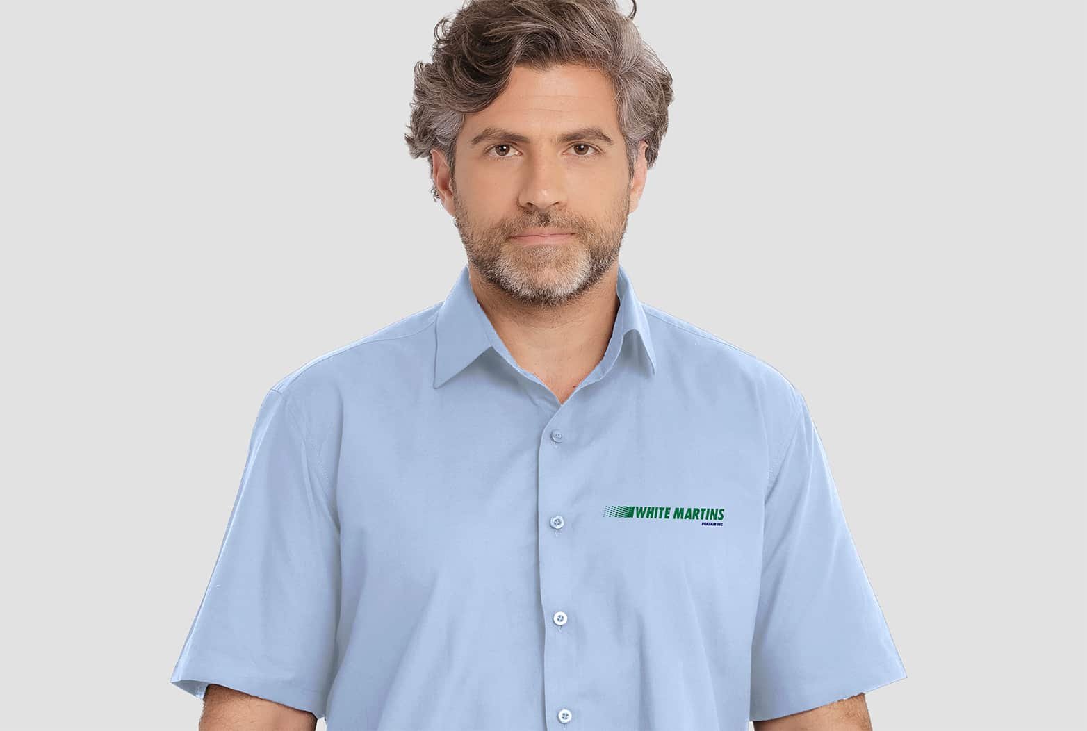 Homem vestindo camisa social masculina azul claro de manga curta com bordado White Martins aplicado no lado esquerdo do peito.