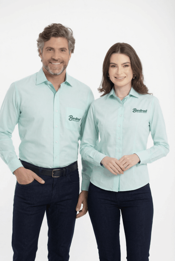 Dois modelos, um masculino e um feminino, em fundo branco vestindo camisas sociais corporativas verde claro personalizadas. A camisa masculina é de manga longa, com colarinho estruturado, bolso frontal no lado esquerdo do peito e bordado com o logo “BemBrasil” em verde escuro aplicado sobre o bolso. A camisa feminina também é de manga longa, com modelagem acinturada, colarinho clássico e bordado “BemBrasil” em verde escuro no lado esquerdo do peito. Ambas as peças apresentam fechamento por botões frontais e excelente caimento, ideais para padronização empresarial.