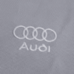 Imagem em close do bordado Audi aplicado em tecido cinza, com os quatro anéis interligados em branco acima da palavra “Audi”, também bordada em branco, evidenciando acabamento preciso e textura do tecido.