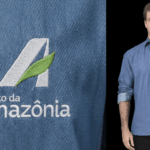 Imagem em close do bordado Banco da Amazônia aplicado em camisa social masculina azul, com símbolo estilizado da letra “A” em branco e detalhe de folha verde, acompanhado do texto “banco da amazônia” em branco. Ao lado, modelo masculino veste a camisa personalizada e aponta para o logotipo no lado esquerdo do peito.