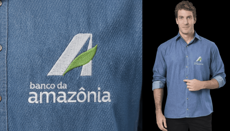 Imagem em close do bordado Banco da Amazônia aplicado em camisa social masculina azul, com símbolo estilizado da letra “A” em branco e detalhe de folha verde, acompanhado do texto “banco da amazônia” em branco. Ao lado, modelo masculino veste a camisa personalizada e aponta para o logotipo no lado esquerdo do peito.