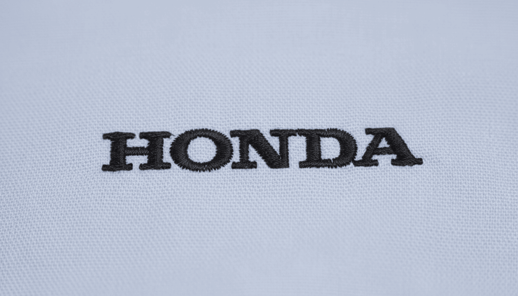 Close-up do bordado “HONDA” em linha preta aplicado sobre tecido azul claro. A imagem destaca a textura do tecido, a densidade dos pontos e o acabamento preciso do bordado institucional, evidenciando a qualidade da personalização.