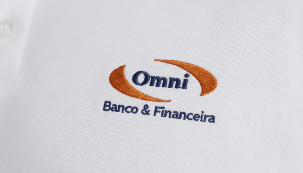 Close-up do bordado “Omni Banco & Financeira” aplicado sobre tecido branco de camisa social corporativa. O logotipo apresenta a palavra “Omni” em azul escuro centralizada entre dois elementos curvos em laranja, formando um símbolo envolvente. Abaixo, a assinatura “Banco & Financeira” aparece bordada em azul. A textura do tecido plano é visível, destacando a qualidade e definição do bordado.