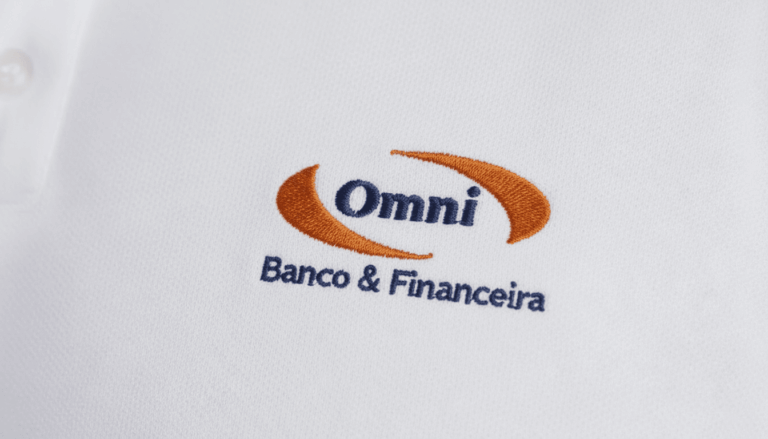 Close-up do bordado “Omni Banco & Financeira” aplicado sobre tecido branco de camisa social corporativa. O logotipo apresenta a palavra “Omni” em azul escuro centralizada entre dois elementos curvos em laranja, formando um símbolo envolvente. Abaixo, a assinatura “Banco & Financeira” aparece bordada em azul. A textura do tecido plano é visível, destacando a qualidade e definição do bordado.
