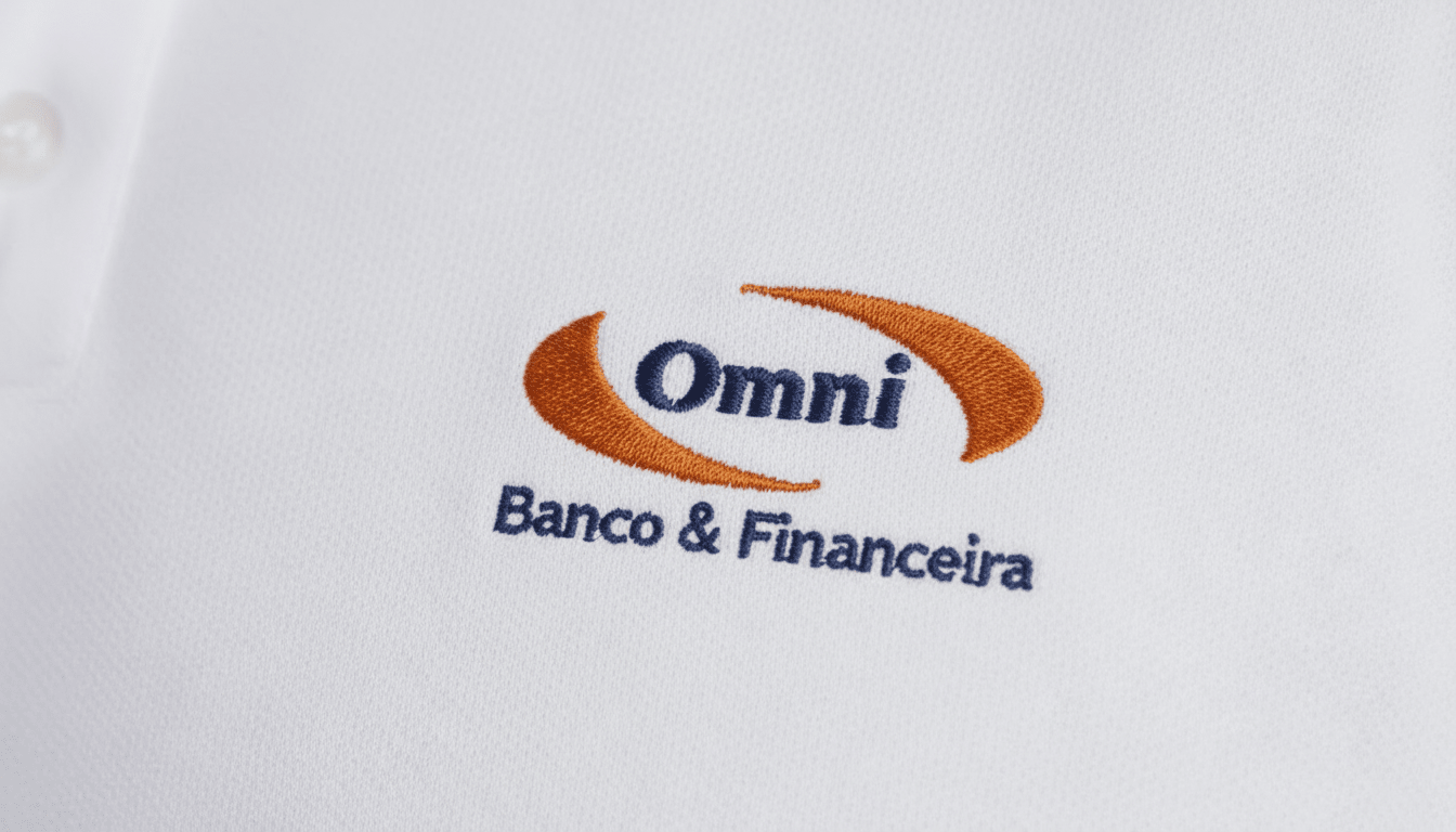 Close-up do bordado “Omni Banco & Financeira” aplicado sobre tecido branco de camisa social corporativa. O logotipo apresenta a palavra “Omni” em azul escuro centralizada entre dois elementos curvos em laranja, formando um símbolo envolvente. Abaixo, a assinatura “Banco & Financeira” aparece bordada em azul. A textura do tecido plano é visível, destacando a qualidade e definição do bordado.