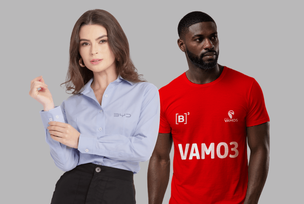 Dois modelos em fundo neutro cinza vestindo uniformes corporativos personalizados. À esquerda, modelo feminina usando camisa social azul clara de manga longa, com colarinho estruturado e fechamento por botões frontais, bordada no lado esquerdo do peito com o logo “BYD” em cinza. À direita, modelo masculino vestindo camiseta vermelha de manga curta com gola careca, personalizada com estampa branca no peito contendo “[B]³” e a palavra “VAMOS”, além de outro logo “VAMOS” menor no lado superior direito do peito. As peças apresentam excelente acabamento e aplicação de identidade visual corporativa.