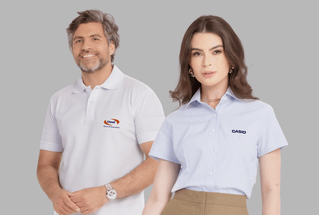 Dois modelos em fundo neutro cinza vestindo uniformes corporativos personalizados. À esquerda, modelo masculino usando polo branca com gola estruturada e fechamento por botões, bordada no lado esquerdo do peito com o logo “Omni Banco & Financeira” em azul e laranja. À direita, modelo feminina vestindo camisa social azul clara de manga curta, com colarinho clássico e fechamento por botões frontais, bordada no lado esquerdo do peito com o logo “CASIO” em azul marinho. As peças apresentam modelagem profissional, excelente caimento e bordado empresarial de alta definição.