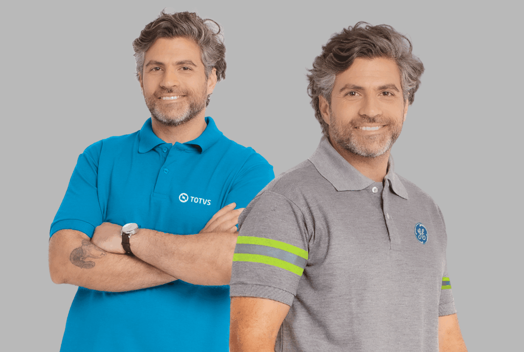 Dois modelos masculinos em fundo neutro cinza vestindo polos corporativas personalizadas. À esquerda, polo azul turquesa com gola estruturada e fechamento por botões, bordada no lado esquerdo do peito com o logo “TOTVS” em branco. O modelo está com os braços cruzados, evidenciando o caimento e a estrutura da peça. À direita, polo cinza mescla com gola clássica e botões frontais, apresentando duas faixas horizontais em verde fluorescente na manga e bordado do logo “GE” em azul no lado esquerdo do peito. Ambas as peças possuem modelagem moderna, acabamento refinado e bordado empresarial de alta definição.