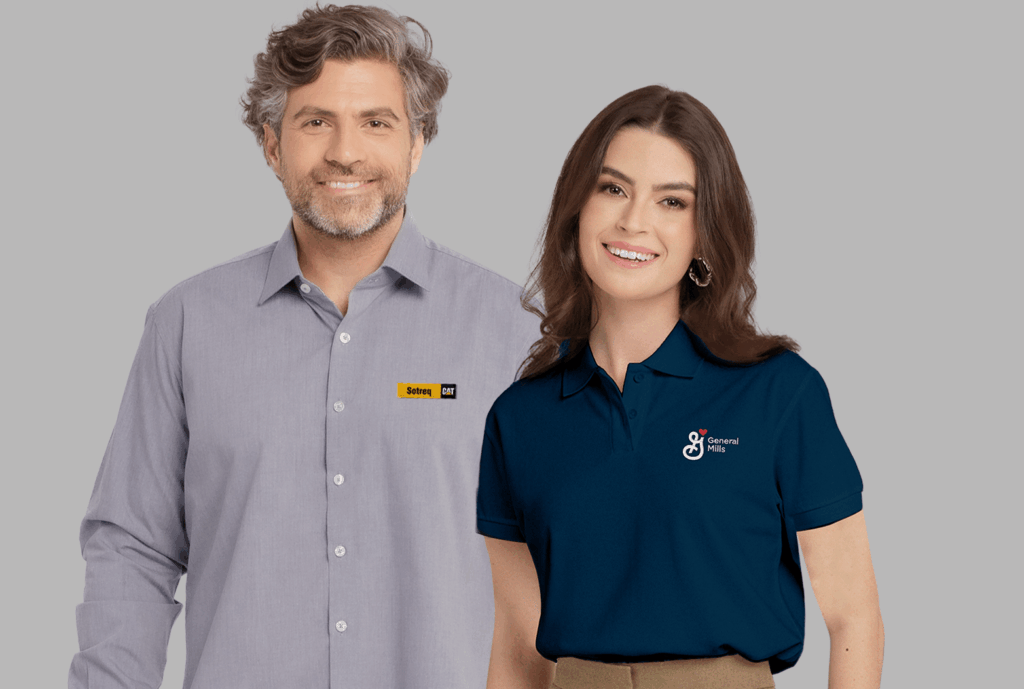 Dois modelos em fundo neutro cinza vestindo uniformes corporativos personalizados. À esquerda, modelo masculino usando camisa social cinza clara de manga longa, com colarinho estruturado e fechamento por botões frontais, bordada no lado esquerdo do peito com o logo “Sotreq CAT” em amarelo e preto. À direita, modelo feminina vestindo polo azul escura com gola estruturada e botões frontais, bordada no lado esquerdo do peito com o logo “General Mills” em branco acompanhado de um pequeno coração vermelho acima da letra “G”. As peças apresentam modelagem profissional, excelente caimento e bordado empresarial de alta definição.