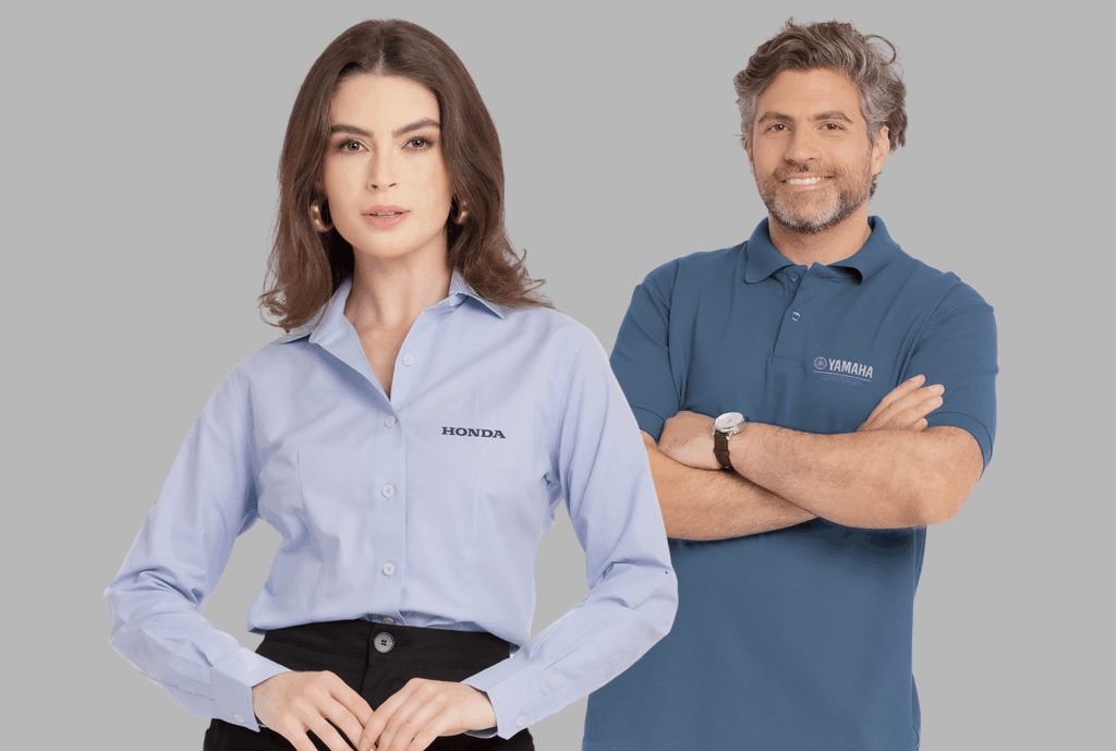 Dois modelos em fundo neutro cinza vestindo uniformes corporativos personalizados. À esquerda, modelo feminina usando camisa social azul clara de manga longa, com colarinho estruturado e fechamento por botões frontais, bordada no lado esquerdo do peito com o logo “HONDA” em preto. À direita, modelo masculino vestindo polo azul escura com gola estruturada e carcela com botões, bordada no lado esquerdo do peito com o logo “Yamaha Consórcio” em branco. As peças apresentam modelagem profissional, excelente caimento e bordado empresarial de alta definição.