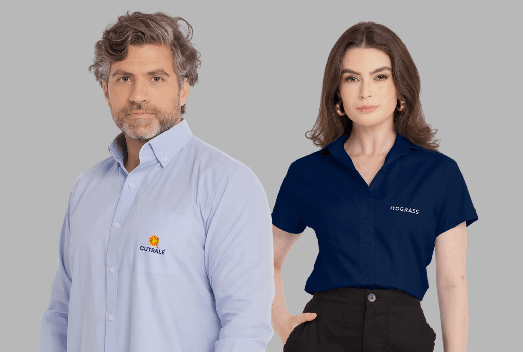 Dois modelos em fundo neutro cinza vestindo uniformes corporativos personalizados. À esquerda, modelo masculino usando camisa social azul clara de manga longa, com colarinho estruturado, bolso frontal no peito esquerdo e fechamento por botões, bordada com o logo “Cutrale” em azul acompanhado de símbolo circular em laranja e amarelo acima do nome. À direita, modelo feminina vestindo camisa social azul marinho de manga curta, com colarinho clássico e botões frontais, bordada no lado esquerdo do peito com o logo “Itograss” em branco. Ambas as peças apresentam modelagem profissional, excelente caimento e bordado empresarial de alta definição.