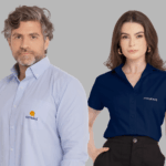 Dois modelos em fundo neutro cinza vestindo uniformes corporativos personalizados. À esquerda, modelo masculino usando camisa social azul clara de manga longa, com colarinho estruturado, bolso frontal no peito esquerdo e fechamento por botões, bordada com o logo “Cutrale” em azul acompanhado de símbolo circular em laranja e amarelo acima do nome. À direita, modelo feminina vestindo camisa social azul marinho de manga curta, com colarinho clássico e botões frontais, bordada no lado esquerdo do peito com o logo “Itograss” em branco. Ambas as peças apresentam modelagem profissional, excelente caimento e bordado empresarial de alta definição.