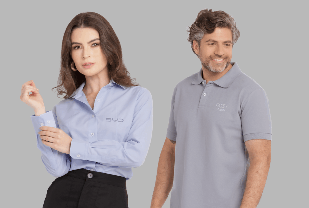 Dois modelos em fundo neutro cinza vestindo uniformes corporativos personalizados. À esquerda, modelo feminina usando camisa social azul clara de manga longa, com colarinho estruturado e fechamento por botões frontais, bordada no lado esquerdo do peito com o logo “BYD” em cinza. A peça apresenta caimento alinhado e acabamento refinado, ideal para ambiente corporativo. À direita, modelo masculino vestindo polo cinza claro com gola estruturada e carcela com botões, bordada no lado esquerdo do peito com o logo “Audi” acompanhado dos quatro anéis característicos da marca em tom discreto. Ambas as peças demonstram padronização empresarial com bordado de alta definição e modelagem profissional.