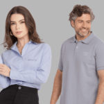 Dois modelos em fundo neutro cinza vestindo uniformes corporativos personalizados. À esquerda, modelo feminina usando camisa social azul clara de manga longa, com colarinho estruturado e fechamento por botões frontais, bordada no lado esquerdo do peito com o logo “BYD” em cinza. A peça apresenta caimento alinhado e acabamento refinado, ideal para ambiente corporativo. À direita, modelo masculino vestindo polo cinza claro com gola estruturada e carcela com botões, bordada no lado esquerdo do peito com o logo “Audi” acompanhado dos quatro anéis característicos da marca em tom discreto. Ambas as peças demonstram padronização empresarial com bordado de alta definição e modelagem profissional.