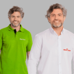 Dois modelos masculinos em fundo neutro cinza vestindo uniformes corporativos personalizados. À esquerda, polo verde vibrante com gola estruturada e carcela com botões escuros, bordada no lado esquerdo do peito com o logo “Nutrien” em branco e detalhe adicional na manga. À direita, camisa social branca de manga longa com fechamento por botões e colarinho estruturado, bordada no lado esquerdo do peito com o logo “Royal Canin” em vermelho. As peças apresentam modelagem profissional, acabamento refinado e bordado de alta definição, ideais para padronização empresarial.