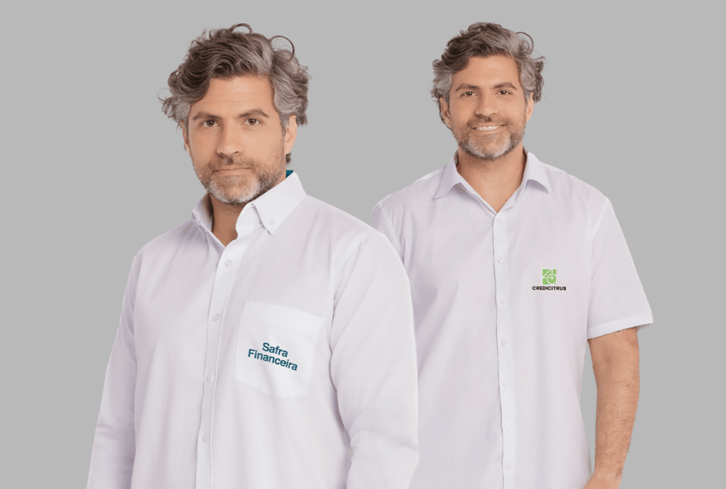 Dois modelos masculinos vestindo camisas sociais corporativas brancas em fundo neutro cinza. À esquerda, camisa social de manga longa com bolso frontal no peito esquerdo e bordado “Safra Financeira” em azul, aplicado sobre o bolso. A camisa possui colarinho estruturado, fechamento frontal por botões brancos e tecido de aspecto leve e profissional. À direita, camisa social branca de manga curta com fechamento por botões e bordado “CREDICITRUS” em preto abaixo de um símbolo verde geométrico, aplicado no lado esquerdo do peito. Ambas as peças apresentam modelagem tradicional e acabamento refinado, ideais para uniformes corporativos personalizados.