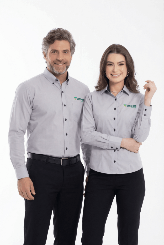 Dois modelos, um masculino e um feminino, em fundo branco vestindo camisas sociais corporativas cinza claro personalizadas. Ambas as camisas possuem manga longa, colarinho estruturado e fechamento por botões pretos que contrastam com o tecido. No lado esquerdo do peito está bordado o logo “SICOOB” em verde, acompanhado do símbolo triangular característico da marca. A camisa masculina apresenta bolso frontal discreto, enquanto a feminina possui modelagem acinturada para melhor ajuste ao corpo. As peças demonstram acabamento refinado e padronização institucional.