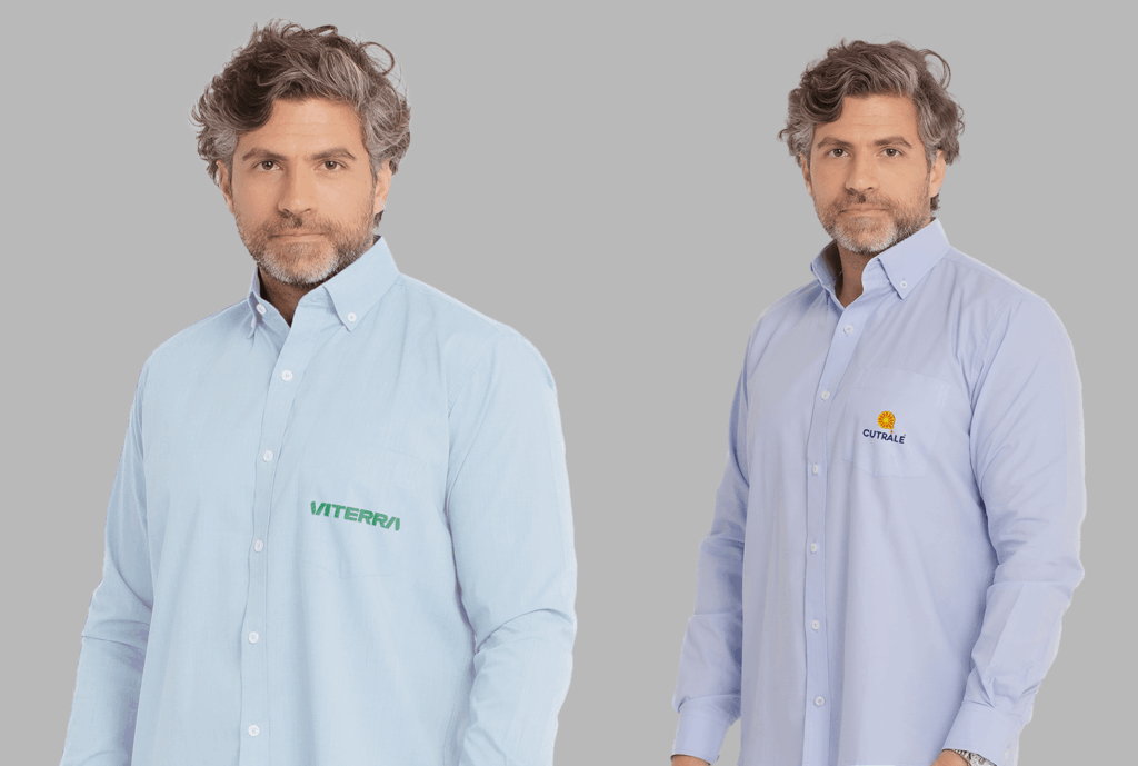 Dois modelos masculinos vestindo camisas sociais corporativas de manga longa em fundo neutro cinza. À esquerda, camisa social azul clara com fechamento por botões brancos e bolso frontal no lado esquerdo do peito, bordada com o logo “VITERRA” em verde. À direita, camisa social azul clara levemente mais intensa, também com botões frontais e bolso no peito esquerdo, bordada com o logo “CUTRALE” em azul marinho acompanhado de um símbolo circular laranja e amarelo acima do nome. As camisas apresentam modelagem tradicional, colarinho estruturado e acabamento refinado, ideais para uniformes corporativos personalizados.