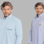 Dois modelos masculinos vestindo camisas sociais corporativas de manga longa em fundo neutro cinza. À esquerda, camisa social azul clara com fechamento por botões brancos e bolso frontal no lado esquerdo do peito, bordada com o logo “VITERRA” em verde. À direita, camisa social azul clara levemente mais intensa, também com botões frontais e bolso no peito esquerdo, bordada com o logo “CUTRALE” em azul marinho acompanhado de um símbolo circular laranja e amarelo acima do nome. As camisas apresentam modelagem tradicional, colarinho estruturado e acabamento refinado, ideais para uniformes corporativos personalizados.