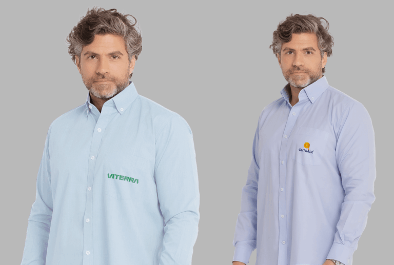 Dois modelos masculinos vestindo camisas sociais corporativas de manga longa em fundo neutro cinza. À esquerda, camisa social azul clara com fechamento por botões brancos e bolso frontal no lado esquerdo do peito, bordada com o logo “VITERRA” em verde. À direita, camisa social azul clara levemente mais intensa, também com botões frontais e bolso no peito esquerdo, bordada com o logo “CUTRALE” em azul marinho acompanhado de um símbolo circular laranja e amarelo acima do nome. As camisas apresentam modelagem tradicional, colarinho estruturado e acabamento refinado, ideais para uniformes corporativos personalizados.