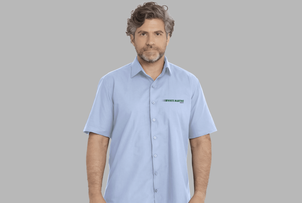 Homem vestindo camisa social masculina azul claro de manga curta com bordado White Martins aplicado no lado esquerdo do peito.