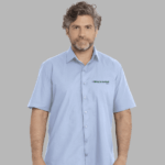 Homem vestindo camisa social masculina azul claro de manga curta com bordado White Martins aplicado no lado esquerdo do peito.