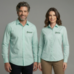 Dois modelos, um homem e uma mulher, vestindo camisas sociais verde-menta personalizadas com o logotipo bordado “Bem Brasil” no lado esquerdo do peito. A camisa masculina possui modelagem reta, mangas longas com punhos abotoados e fechamento frontal por botões. A camisa feminina apresenta modelagem acinturada, mangas longas e fechamento por botões frontais, valorizando o caimento corporativo. Ambas as peças estão bem passadas, com acabamento refinado e tecido de aspecto leve, em fundo neutro cinza claro.