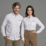 Dois modelos, um homem e uma mulher, vestindo camisas sociais brancas personalizadas com o logotipo bordado “NISSAN DAHRUJ” no lado esquerdo do peito em vermelho. A camisa masculina possui modelagem reta, mangas longas com punhos abotoados, bolso frontal e fechamento por botões com detalhe interno em tom avermelhado. A camisa feminina apresenta modelagem levemente acinturada, mangas longas, bolso frontal e fechamento por botões com detalhe contrastante, proporcionando caimento elegante e alinhado. As peças têm tecido liso, aparência sofisticada e acabamento profissional, posicionadas em fundo neutro cinza claro.