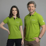 Dois modelos, uma mulher e um homem, vestindo polos verde vibrante personalizadas com o logotipo bordado “Nutrien” no lado esquerdo do peito em branco. A polo feminina possui modelagem levemente acinturada, mangas curtas ajustadas e gola polo com botões frontais em contraste escuro na parte interna. A polo masculina apresenta modelagem reta, mangas curtas e gola estruturada com fechamento por botões. Ambas as peças exibem também detalhe de logotipo aplicado na manga. O tecido tem aparência confortável e resistente, ideal para uso corporativo, em fundo neutro cinza claro.